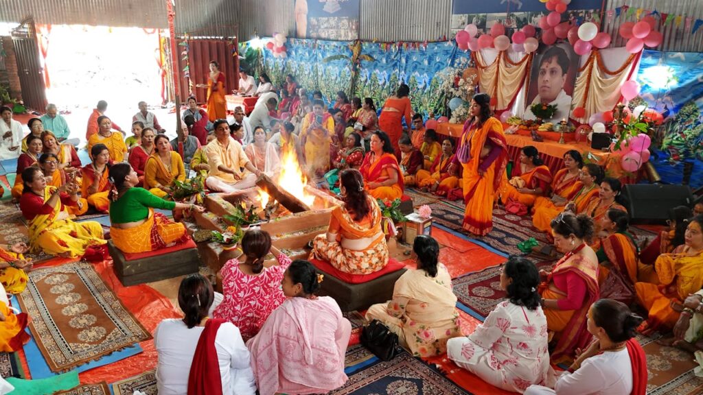 A vibrant indoor Hindu religious ceremony with participants in colorful traditional attire surrounding a sacred fire.